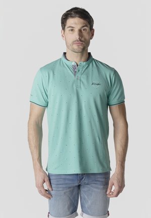 Hombre con camiseta tipo polo de color verde menta con estampado y cuello abotonado, y pantalones cortos de mezclilla azul claro, posando contra un fondo liso.