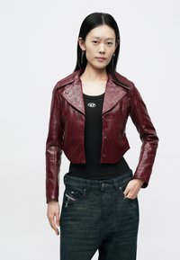 Bordeauxfarbene, kurz geschnittene Lederjacke mit weitem Kragen und Druckknopfverschluss, kombiniert mit einem schwarzen Tanktop und dunkelblauen Jeans.