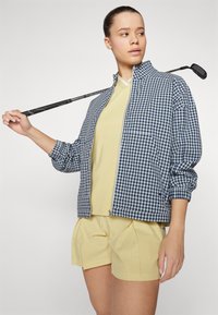 Σακάκι σε ναυτικό και λευκό καρό με κλείσιμο φερμουάρ, συνδυασμένο με ανοιχτό κίτρινο μπλουζάκι και πλισέ σορτς. Το μοντέλο κρατάει ένα γκολφ κλαμπ πάνω στον ώμο της.