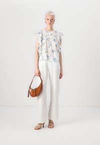 Blusa bianca trasparente con motivo floreale e maniche a ruffle, abbinata a pantaloni larghi in lino bianco e una borsa in pelle stampata coccodrillo marrone.