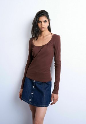 Jeune femme aux cheveux foncés portant un haut marron à manches longues et une jupe en jean bleue boutonnée, posant devant un fond blanc.