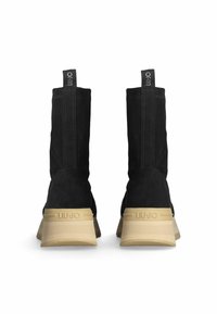 Stivaletti neri in suede con design alto, suole in gomma beige chiaro e linguette sul tacco con marchio. Texture liscia e estetica minimalista.