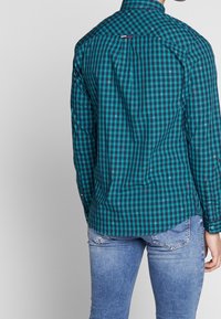 Camicia a maniche lunghe a quadri turchesi e neri con colletto a punta e dettaglio sulla spalla dietro, indossata con jeans blu chiaro.