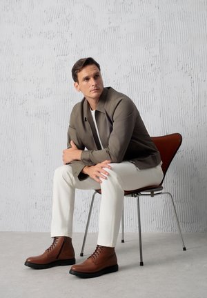 Brązowe skórzane buty z sznurowaniem na czarnej podeszwie, noszone z białymi spodniami i dopasowaną szaro-skalną marynarką, siedzące na krześle z czerwonymi akcentami.