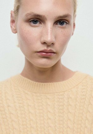 Joven con piel clara, ojos azules, pecas y cabello rubio que lleva un suéter de punto trenzado color crema frente a un fondo liso.