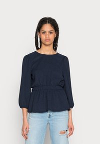 Top peplum azul-marinho com mangas longas e bufantes e cintura elástica, combinado com jeans de denim claro com detalhes desgastados.