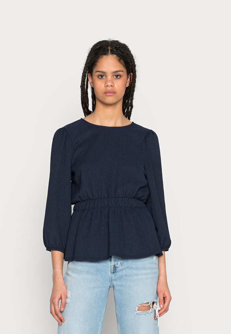 Top peplum azul-marinho com mangas longas e bufantes e cintura elástica, combinado com jeans de denim claro com detalhes desgastados.