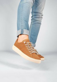 Zapatillas de ante marrón con suela de goma crema, que presentan cordones planos beige y un logo en la lengüeta. Llevadas con jeans claros azules remangados.