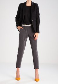 Černý blazer přes černý top, doplněný šedými šitými kalhotami a žlutými podpatky. Bílý pásek dodává kontrast. Hladká textura, přiléhavý design.