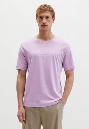 Junger Mann mit blondem Haar, der ein hellviolettes Marc O’ Polo T-Shirt und beige Hosen trägt, steht vor einem schlichten weißen Hintergrund.