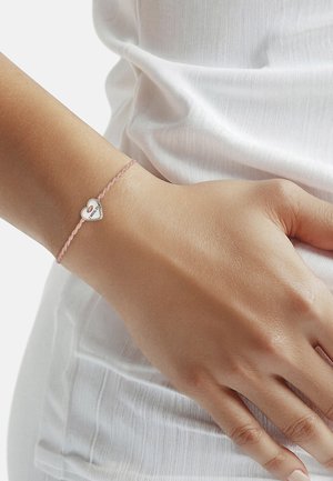 Pols met een lichtroze gevlochten armband met een zilveren hartbedel met gegraveerde tekst, gedragen over een wit, gestructureerd stoffen top.