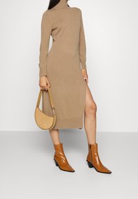 Strickkleid mit langen Ärmeln und Rollkragen in Beige mit einem seitlichen Schlitz. Kombiniert mit spitzen, tanfarbenen Stiefeletten und einer kleinen, geschwungenen Handtasche.