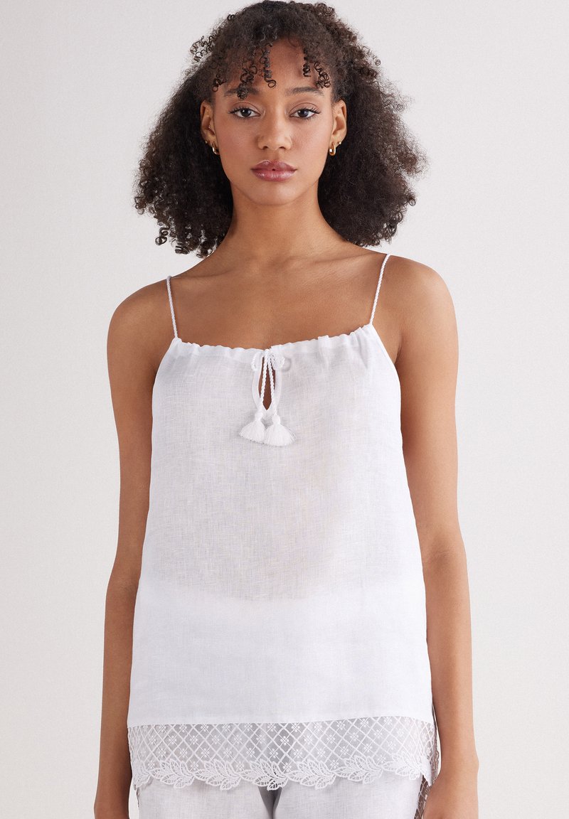 Witte linnen camisole met verstelbare spaghettibandjes, een ronde halslijn met kwastaccenten en een kanten rand aan de onderkant.
