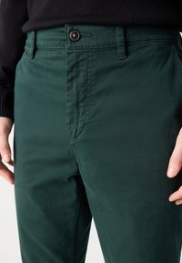 Pantalon vert foncé avec fermeture à bouton noir porté par une personne en pull noir, mains reposant près des hanches.