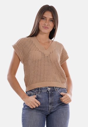 Donna che indossa un maglione beige senza maniche in maglia ad uncinetto con scollo a V e jeans blu a vita alta, mani nelle tasche.
