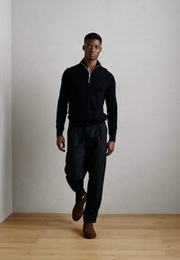 Zwarte gebreide trui met rits en zwarte broek, beide van een gladde textuur. Het model draagt bruine schoenen met een minimalistisch ontwerp tegen een neutrale achtergrond.
