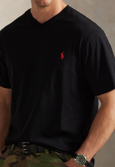 Άνδρας που φοράει μαύρο T-shirt με V λαιμόκοψη και κόκκινο λογότυπο, παντελόνι παραλλαγής, μαύρη ζώνη και ρολόι χειρός με πράσινο καντράν, στέκεται με το ένα χέρι στην τσέπη.