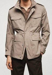 Veste légère beige avec col montant, deux poches poitrine, taille élastique et fermetures à boutons. Texture lisse, design décontracté.