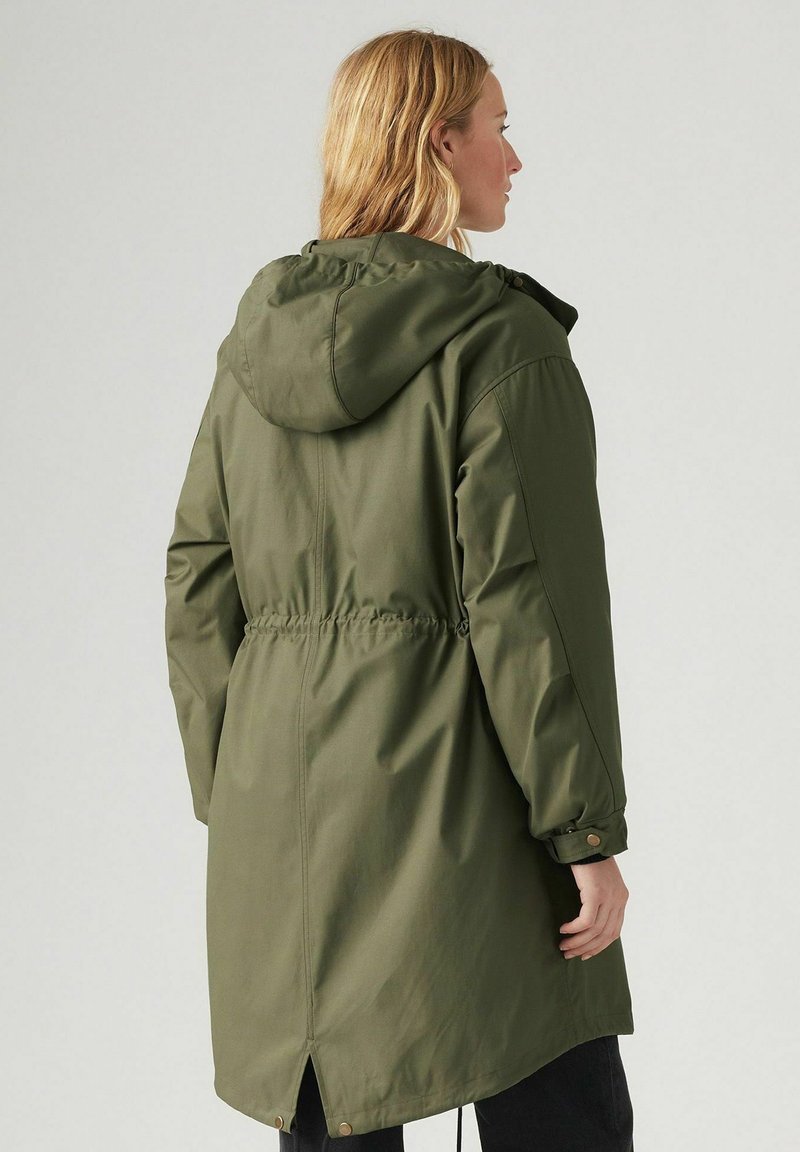 Donna che indossa un lungo parka verde oliva con cappuccio, vita cintrata, polsini abbottonati e spacco posteriore, di profilo contro uno sfondo neutro.