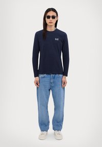 Mornarska bluza s dugim rukavima s logotipom, svijetloplave široke traperice i bijele tenisice. Dodatna oprema uključuje tamne sunčane naočale. Jednostavan, ležeran outfit.