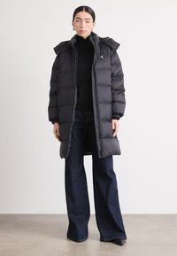 Svart puffjakke med hette, med en quiltet design, lang lengde og elastiske mansjetter. Brukt over en svart høyhalset genser og mørke jeans.