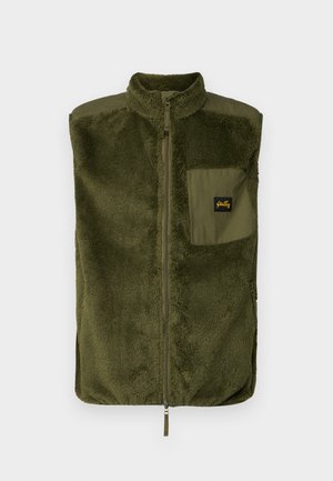 Fleece vest in dark olive green with high collar, front zipper, and a left chest pocket featuring a logo patch. Texture is soft and plush.