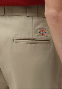 Beige Dickies pants featuring a back pocket and logo patch. Made of textured fabric with a clean, tailored fit and belt loops.