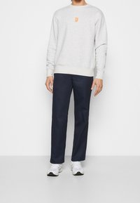 Szara bluza z pomarańczowym logo na klatce piersiowej, w połączeniu z ciemnogranatowymi spodniami i białymi trampkami. Prosty design, luźny krój i casualowy styl.