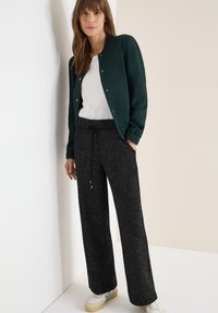 Bomber verde scuro con bottoni a pressione, t-shirt bianca e pantaloni neri a gamba larga con tessitura. Sneakers casual con dettagli crema e blu.