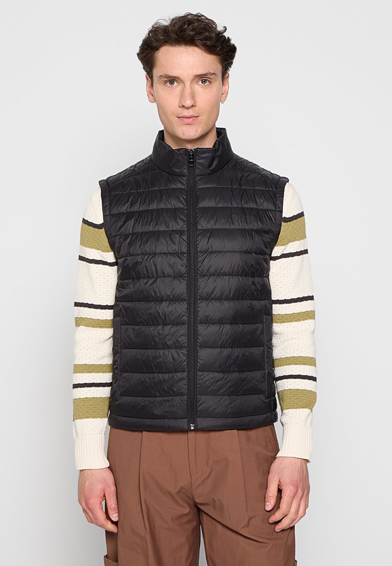 Gilet imbottito nero con zip frontale, dotato di un colletto alto e texture trapuntata. Indossato sopra un maglione a righe e abbinato a pantaloni larghi.