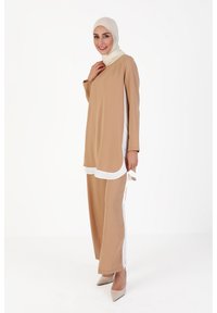 Tunique beige avec des panneaux latéraux blancs et des manches longues, assortie à un pantalon beige. Modèle portant un hijab de couleur claire et des talons.
