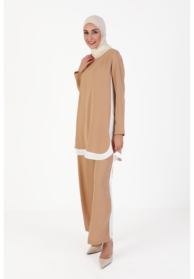 Beige tuniek met witte zijpanelen en lange mouwen, gecombineerd met bijpassende beige broeken. Model draagt een lichtgekleurde hijab en hakken.