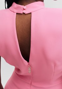 Rückansicht einer Person in einem rosa Kleid mit hohem Kragen, zwei Knöpfen und einem offenen Rücken, der mit einem Reißverschluss mit "HUGO"-Charm gesichert ist.