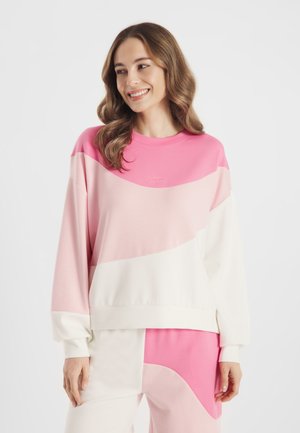 Lachende vrouw met golvend bruin haar, gekleed in een losse sweater met kleurvlakken in roze, lichtroze en wit, en bijpassende broek tegen een witte achtergrond.