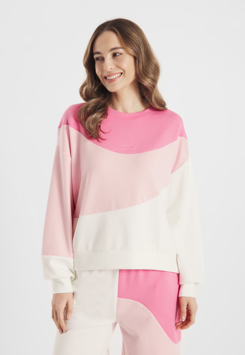 Lachende vrouw met golvend bruin haar, gekleed in een losse sweater met kleurvlakken in roze, lichtroze en wit, en bijpassende broek tegen een witte achtergrond.