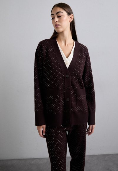 Bordeaux vest met een V-hals, knoopsluiting, twee voorzakken en een wit stippenpatroon. Gdragen over een wit blouse. Gebreide textuur.
