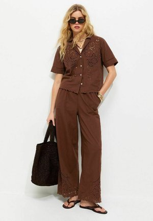 Kobieta ubrana w brązową, krótko rękawową haftowaną koszulę i pasujące szerokie spodnie, niosąca czarną plecioną torbę, w okularach przeciwsłonecznych i sandałach.