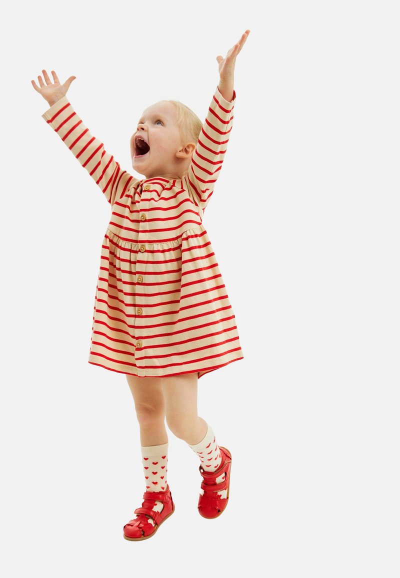 Punane ja kreemjastriibuline kleit nupudega, koos punaste kingade ja väikeste punaste südametega valgete sokkidega. Laps käed üles tõstetud põnevusest.