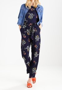 Granatowy kombinezon w kwiaty z wiązanym pasem, połączony z dżinsową kurtką i pomarańczowymi klapkami. Wzór kwiatowy w różnych kolorach.