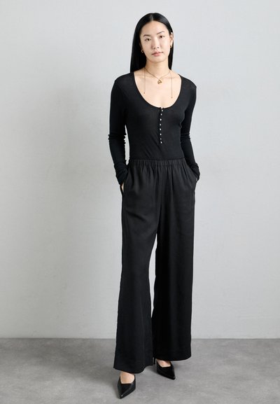 Black long-sleeve bodysuit with buttons at the front, paired with high-waisted, wide-leg black trousers. Pointed black heels.