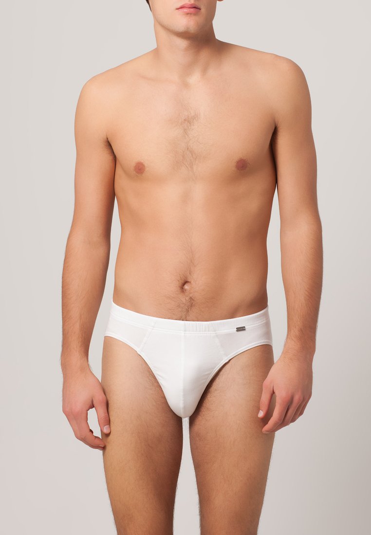 Slip bianco in cotone con una consistenza liscia, design aderente e un sottile logo sulla vita. Modello in piedi, braccia lungo i fianchi.