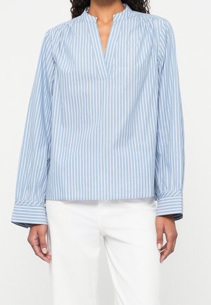 Femme portant un chemisier à rayures verticales bleues et blanches à manches longues avec un col en V fendu et un pantalon blanc, debout devant un fond uni.