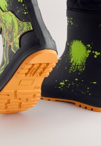 Stivali neri in gomma con motivo a spruzzi verde brillante, suola arancione texturizzata per una migliore aderenza, e parte superiore decorata con una grafica di dinosauro.
