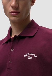 Burgundowa koszulka polo z teksturowanej tkaniny z białym haftowanym logo "WRSTBHVR 030" na lewej piersi oraz dwoma guzikami.