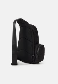 Svart slyngetaske med et teksturert meshpanel, glatt stoff, glidelommer og justerbar stropp. Har et kompakt, asymmetrisk design.