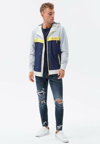 Bunda z větruodolného materiálu v námořnické modré a světle šedé barvě s žlutým pruhem. Slim-fit džíny s odřenými detaily a bílé tenisky.