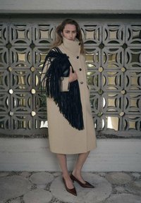 Beżowy płaszcz z wysokim kołnierzem i czarnymi guzikami, ozdobiony czarną torebką z frędzlami. Modelka ma na sobie brązowe, spiczaste baleriny i stoi na tle teksturowanej scenerii.