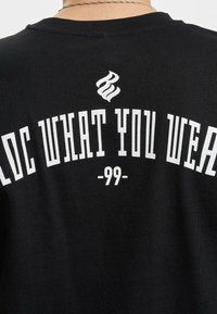Black cotton T-shirt featuring white text that reads "ROC WHAT YOU WEAR" and "-99-", with a stylized graphic above. Text is bold and prominent.