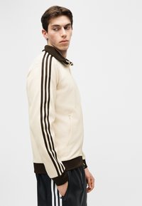 Mladý muž nosiaci béžovohnedú bundu na zips s čiernym dizajnom troch prúžkov na rukávoch, pozerajúci sa doľava s rukami v kapsách.