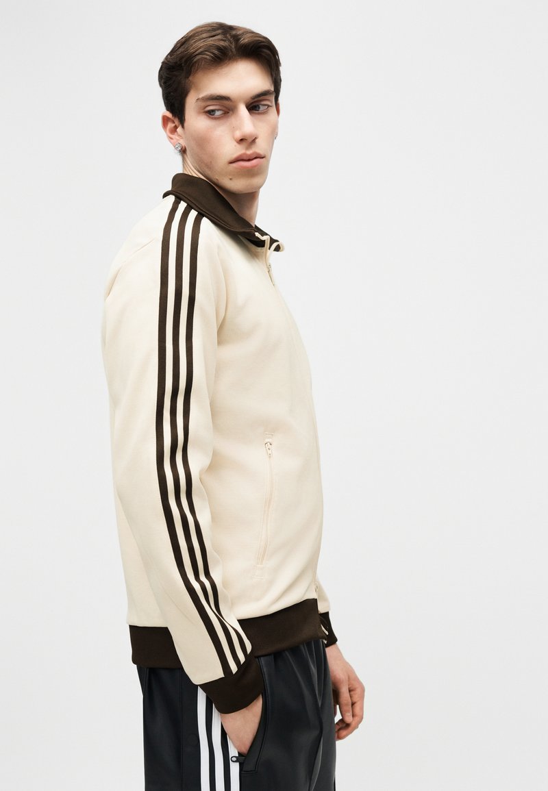 Joven con chaqueta con cremallera beige y marrón con un diseño de tres rayas negras en las mangas, mirando hacia su izquierda con las manos en los bolsillos.